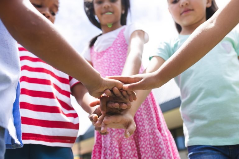 In school outside, diverse group of young students are joining hands. They are wearing casual clothes and enjoying a sunny day outside, unaltered